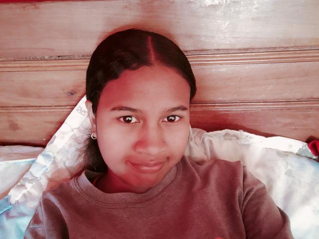 LoderFist, a brown-skinned girl with black hair in a ponytail, wearing a brown shirt, smiling slightly, sitting against a wooden headboard with a patterned pillow.