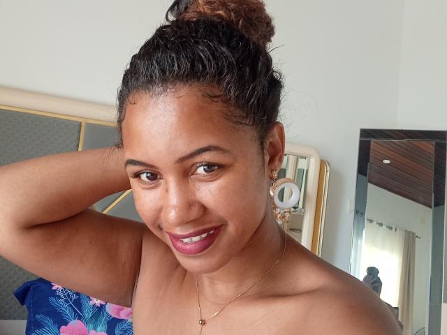 LayahCoquine, with brown skin and curly hair in a bun, smiles at the camera, showing her pink lipstick and gold hoop earrings, with her arm raised, and no clothes.