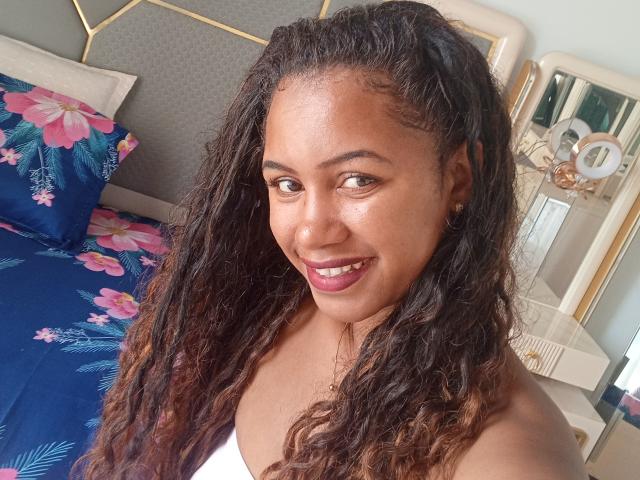 LayahCoquine, a Black woman with curly, dark brown hair, smiles at the camera. She has medium brown skin, dark brown eyes, and wears a white tank top. Her lips are painted pink. The background includes a blue floral bedspread, a grey headboard, and a mirror with circular decorations. The image is a close-up selfie. Her hair is textured and slightly wavy, with a few lighter brown highlights. She has small, gold earrings.