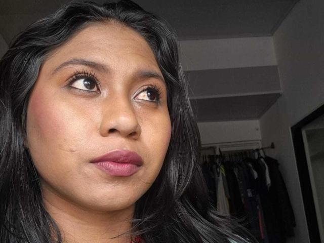 EbonyxLust a la peau brune, les cheveux noirs et ondulés, les yeux marrons, du rouge à lèvres rose et une légère moue. Ses seins sont petits et elle regarde sur le côté..