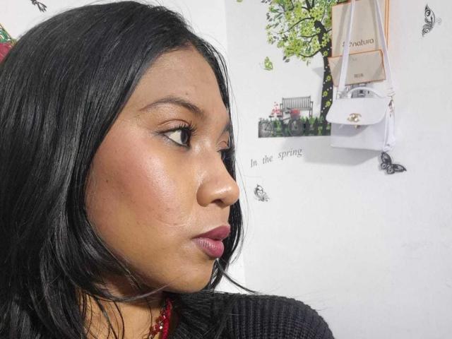 EbonyxLust's face in a photo, dark straight hair, brown skin, subtle makeup, pink lipstick, looking to the side, red necklace, black top, white wall with butterfly decals.