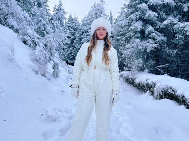 YaseminSaf standing in a snowy forest, wearing a white puffy coat, matching hat, gloves, and belted pants, with long brown hair and a subtle smile.
