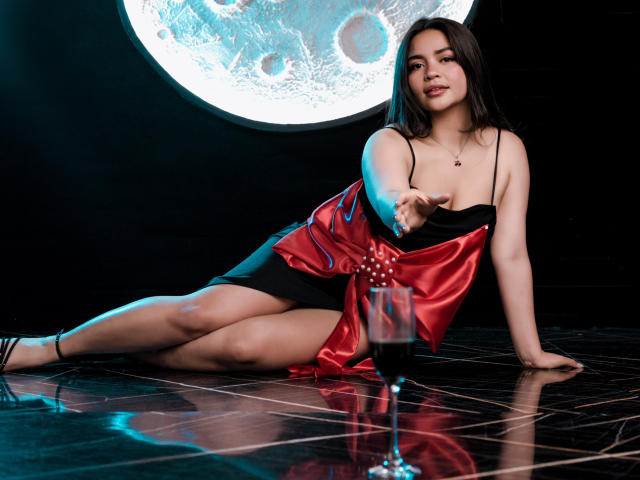 KimDevinne, avec sa poitrine visible, portant une robe en satin rouge et noir, allongée sur des carreaux noirs, tendant la main vers un verre de vin rouge, avec la lune en arrière-plan, et une allure séduisante..