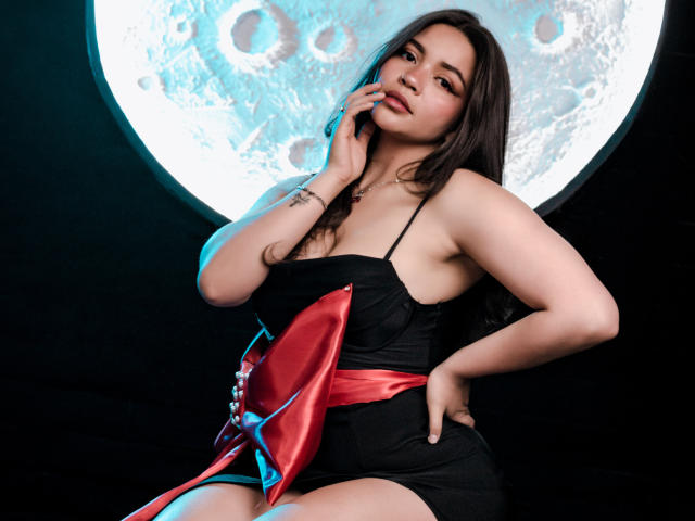 KimDevinne with dark hair, wearing a black dress with red and blue satin accents, touching her cheek, hand on hip, moon in background, cute and sexy.