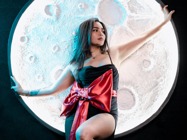 KimDevinne with long dark hair, wearing a black strapless dress with a big red bow, arms raised, standing in front of a glowing circular moon-like light. Her tits are visible.