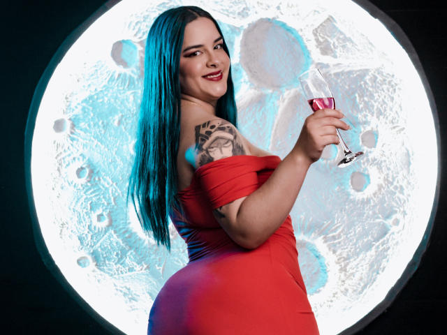 AuroraSantorini with blue hair, red off-shoulder dress, holding a wine glass, smiling at the camera, showing her tattooed arm and round ass. She's standing with a sexy look.