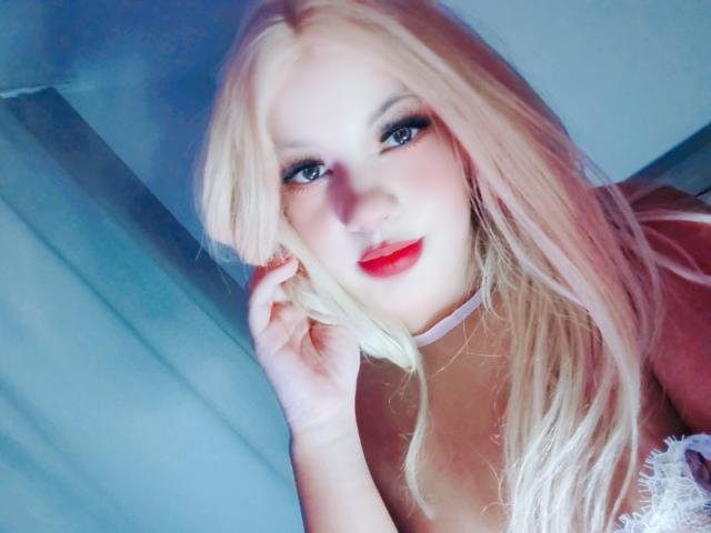 RoseBellee has blonde hair, red lipstick, blue eyes, and a white choker. She's touching her hair with a slight smile, showing her tits in a lace top.