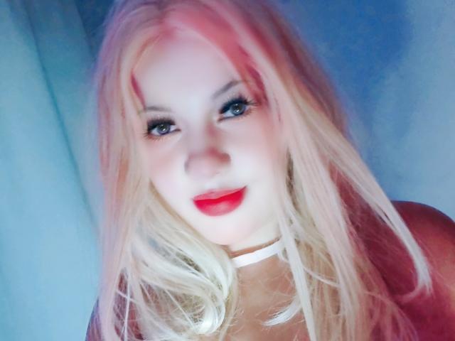 RoseBellee has blonde hair with pink highlights, blue eyes, red lipstick, and a white choker. Her face is slightly blurred, giving a soft, dreamy effect.