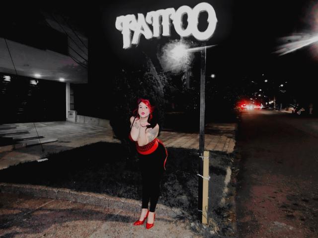 Alexandrapsique with red hair and red heels, wearing a black top and red belt, standing in front of a Pantoo sign at night, holding her tits with a smile.