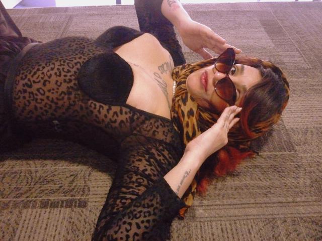 Alexandrapsique lies on a carpet, wearing a sheer black leopard-print dress, black bra, and brown glasses, with a leopard scarf and red-tipped brown hair. Her tits are visible.