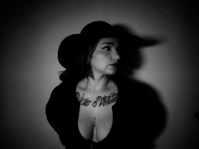 Black-and-white photo of Alexandrapsique with medium tits, wearing a hat, topless, showing a chest tattoo, and a nipple piercing, gazing to the side, shadowed.