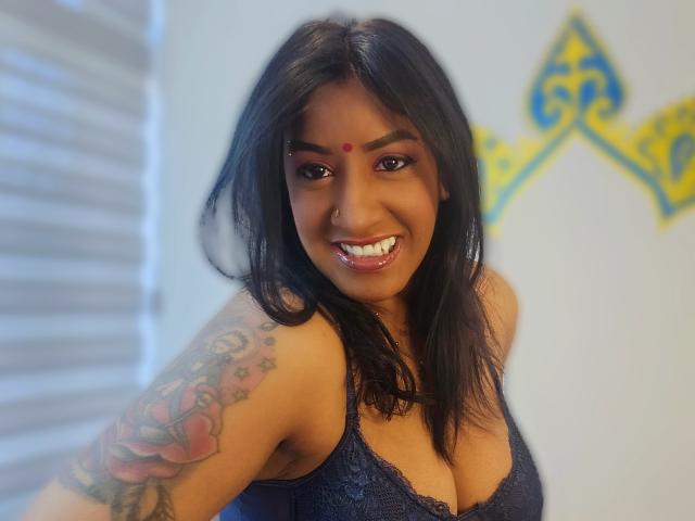 Nahirr with dark hair, smiling, wearing a blue lace bra, showing big boobs, nose ring, red dot on forehead, and a colorful tattoo on her right arm.