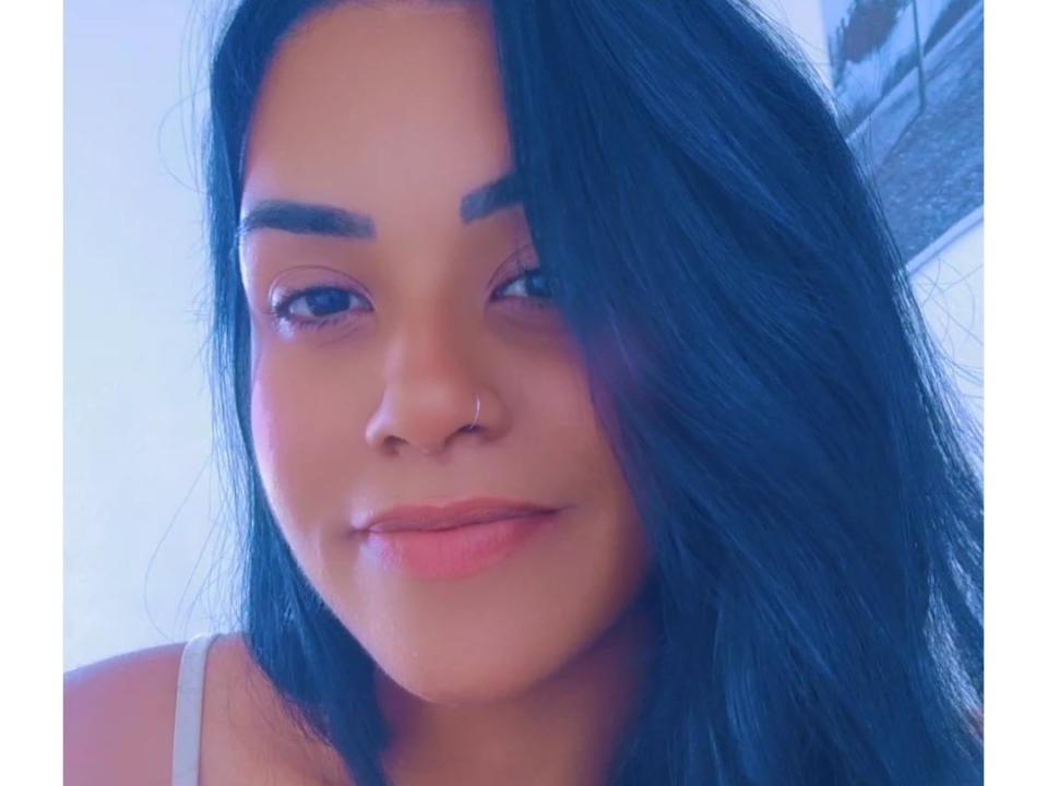 Morenaps, com cabelo azul-escuro, sobrancelhas castanhas, piercing no nariz e lábios rosados, sorrindo suavemente, vestindo uma blusa branca, olhos azuis, pele clara e um leve rubor rosado nas bochechas..