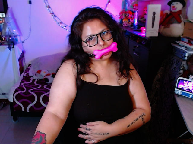 LilithHeavy with black tank top, glasses, pink bone gag, wavy dark hair, tattoos on arm, holding belly, sitting in room with colorful lights, phone on table.