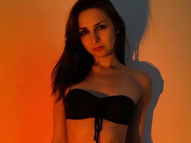 RuthPark with long brown hair, wearing a black strapless bikini top, showing her tits, standing in warm orange and blue lighting, shadow on the wall.