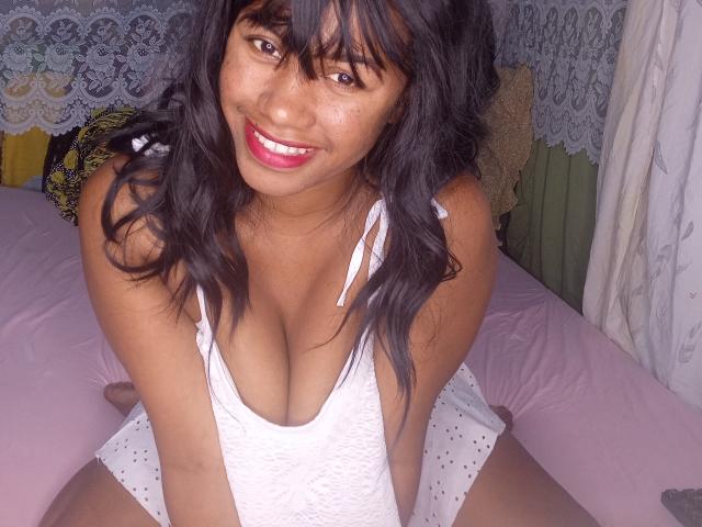 LayahCoquine with dark wavy hair, smiling, wearing a white lace tank top, showing her big tits, sitting on a pink bed, pink lips, brown eyes, cute, attractive.