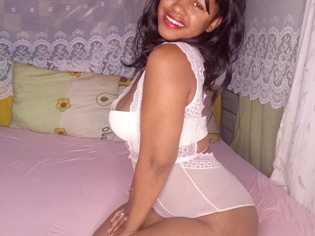 LayahCoquine, smiling, in white lace lingerie, showing her big tits and curvy ass, kneeling on a pink bed, dark brown skin, black wavy hair, red lipstick.