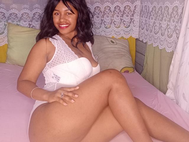 LayahCoquine with dark wavy hair, smiling, wearing a white lace dress, sitting on a pink bed, showing her thick thighs and small boobs, with her hand on her ass.