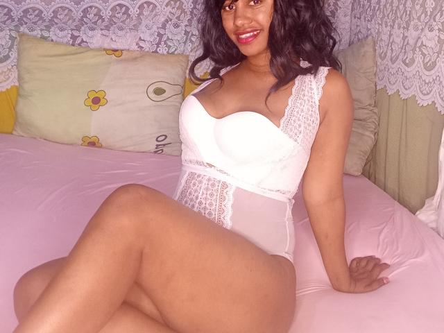 LayahCoquine sits on a pink bed, wearing a white lace and mesh bodysuit. She has dark curly hair and a warm smile. Her legs are crossed, and she's leaning back with her right hand on the bed. The bodysuit has lace details on the shoulders and chest. The background includes a lace curtain and a pillow with yellow flowers. The lighting is soft, highlighting her brown skin and the texture of the lace.