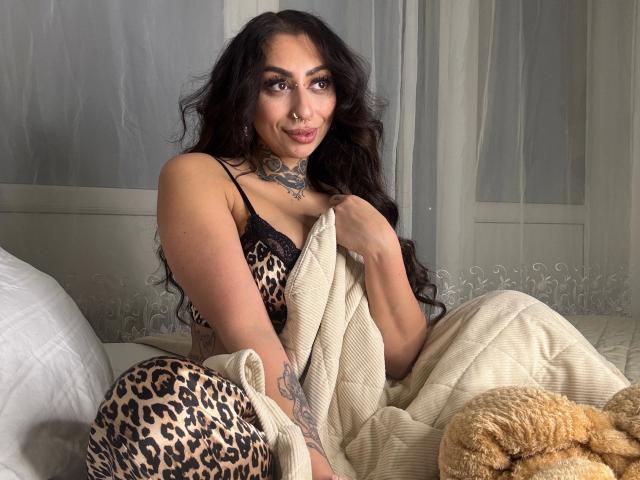Pinkerbell, con cabello largo y negro, un vestido de estampado de leopardo y tatuajes, sosteniendo una manta beige, sentada en una cama con un peluche color arena. Sus pechos están parcialmente cubiertos..
