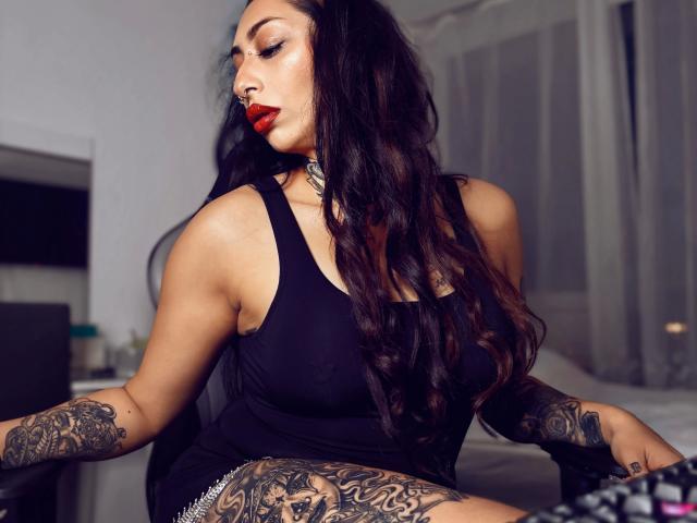 Pinkerbell, com aparência sensual, cabelos longos e escuros, batom vermelho, top preto e tatuagens, sentada com os braços abertos, exibindo tatuagens nos braços e coxas, usando um colar..
