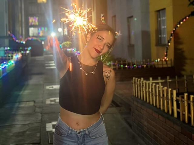 AliceHotBlue with sparkler in hand, wearing black crop top, blue jeans, red lipstick, and necklace, standing on street at night, visible tattoo on right shoulder, smiling.