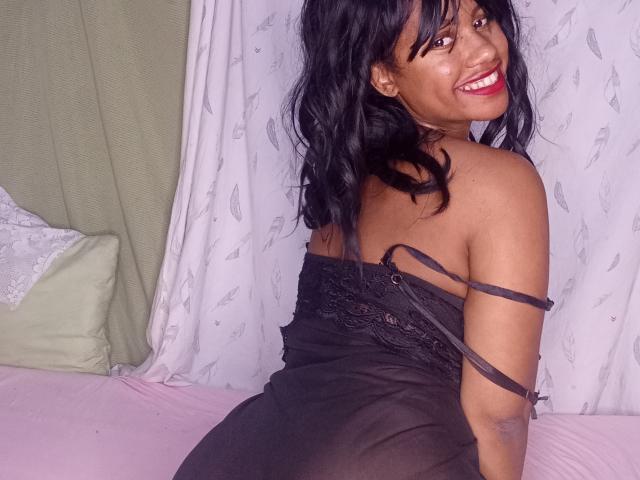 LayahCoquine has dark, wavy hair and a bright red lipstick smile. She's sitting on a pink bed, wearing a sheer black lace dress with straps slipping off her shoulders. Her brown skin contrasts with the black dress and pink bed. The background features white curtains with a subtle feather pattern. The image is clear and focuses on her smiling face and upper body. She has a curvy figure and is looking over her shoulder.