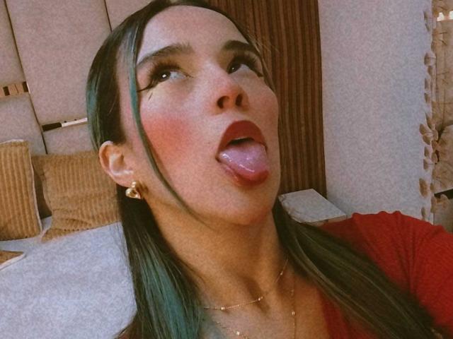 AliceHotBlue with green-tinted hair, tongue out, red lips, and gold earrings, wearing a red top, looking up with brown eyes, and a slight blush on her cheeks.