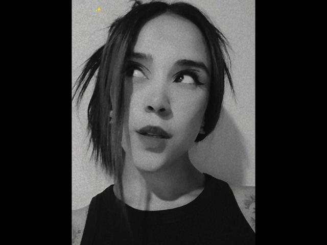 AliceHotBlue has short, messy black hair, looking upward with a slightly open mouth. She's wearing a black shirt, and the grayscale photo has a subtle yellow dot.