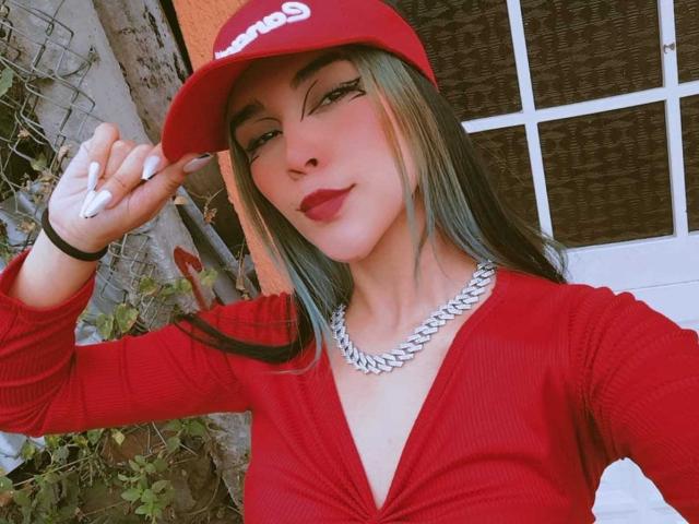 AliceHotBlue with blue-green hair, red lipstick, red cap, red cardigan, silver necklace, and white nails, holding a cigarette, showing her tits, standing outside.