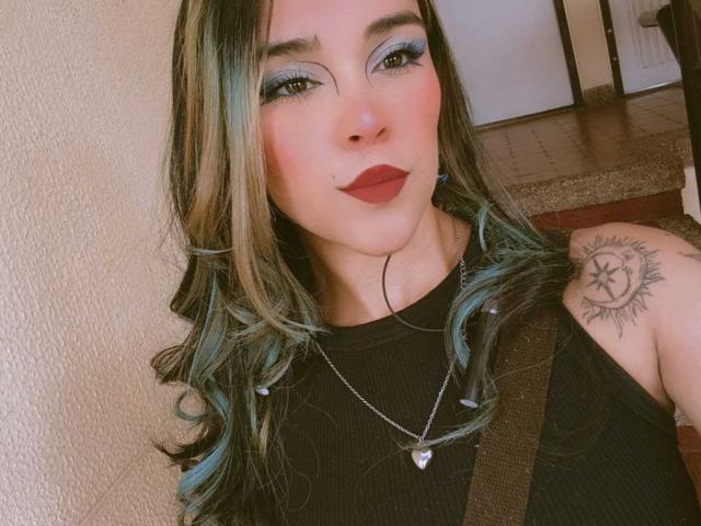 AliceHotBlue with blue-tipped hair, black sleeveless top, red lipstick, blue eyeshadow, tattoo on right shoulder, necklace, strap over shoulder, smiling, indoors.
