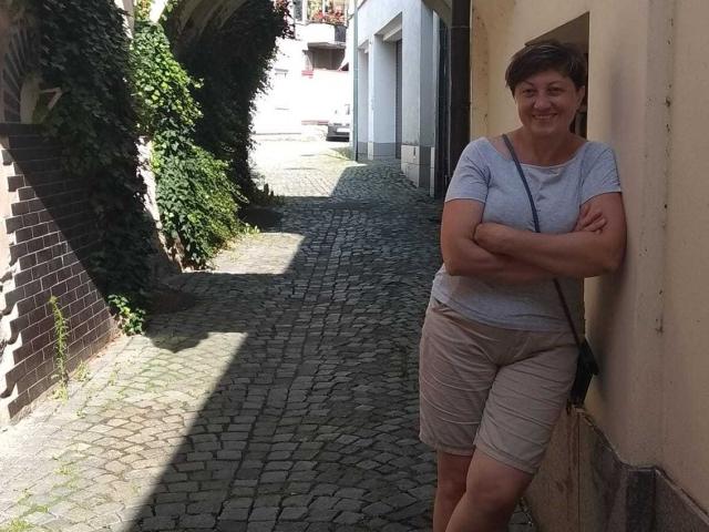 EmilyLlove stands with arms crossed, wearing a gray shirt and beige shorts, leaning against a wall on a cobblestone path, smiling. Short brown hair, small breasts, medium build.