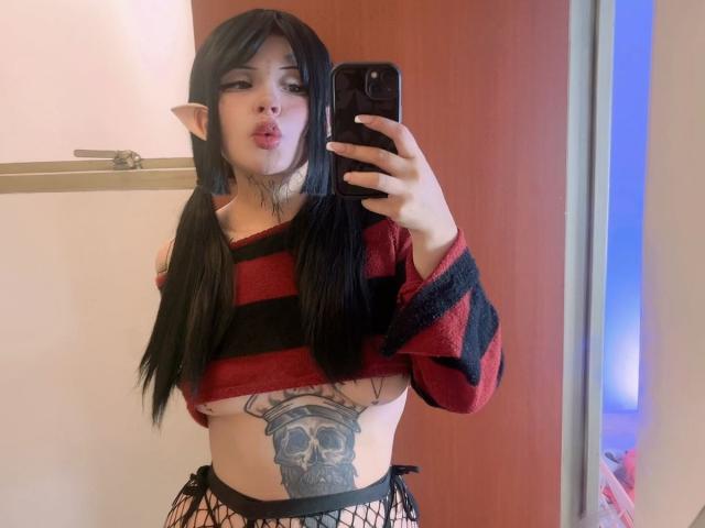 StarWitch with long black hair, elf ears, pouty lips, red and black striped top, showing a skull tattoo on her flat, bare tits, and fishnet bottoms, taking a selfie.