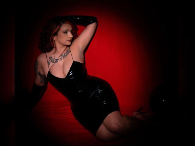 Alexandrapsique with red hair, black latex dress, and gloves, tattooed, leaning seductively on red-lit floor, showing her tits and ass, looking sultry.