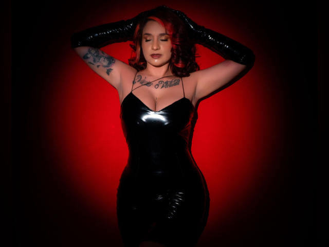 Alexandrapsique in a shiny black latex dress, black gloves, red background, eyes closed, arms up, tattoo on right arm, wavy red hair, full tits, curvy, confident, sexy.