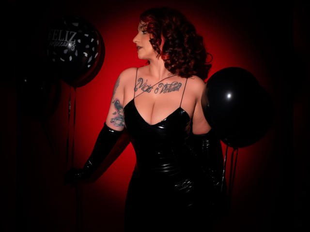 Alexandrapsique with curly red hair, black latex dress, and gloves, holding black balloons, has tattoos on chest and arm, and large boobs, standing against a red-lit background.