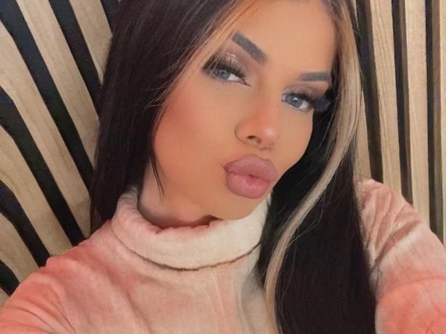IvyRaven with pouty pink lips, blue eyes, thick eyelashes, and blonde-streaked black hair, wearing a pink turtleneck. Boobs not visible. Background is blurred.