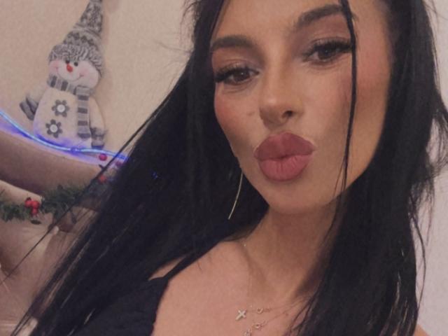 KinkyAmelie with long black hair, pouty pink lips, and thick eyelashes, wearing a black top, gold hoop earrings, and a delicate necklace, making a kissy face.