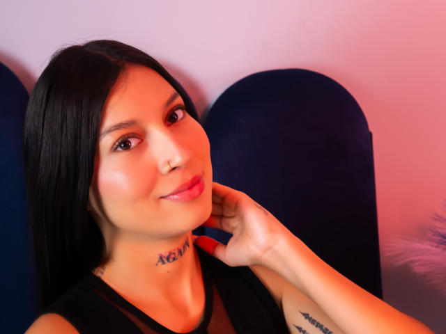 CloeThonsom with long black hair, nose ring, pink lipstick, tattoos on neck and arm, hand on cheek, smiling, wearing black top, blue chair, pink and orange lighting.