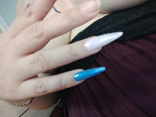 AlinaMistress's hand with long, white and blue glitter nails, showing off her nails against a black and maroon dress. Boobs visible in the background.