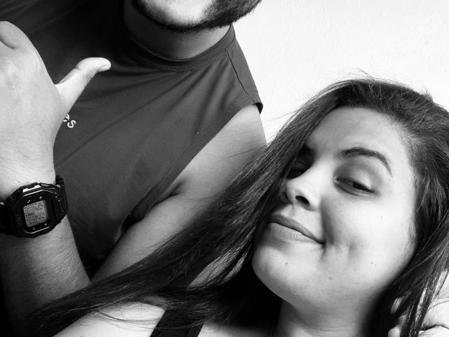 Black and white photo of Casalsativa. Casalsativa, a woman with long, straight hair, rests her head on a man's shoulder. She has a small smile, her eyes half-closed. The man, partially visible, has a beard and wears a sleeveless shirt with a watch on his left wrist. His right arm is around Casalsativa. The man's skin is slightly darker than Casalsativa's. The image focuses on their upper bodies and faces. The woman's hair is dark and smooth. The man's wristwatch is square with a textured face. The photo has a candid, intimate feel.