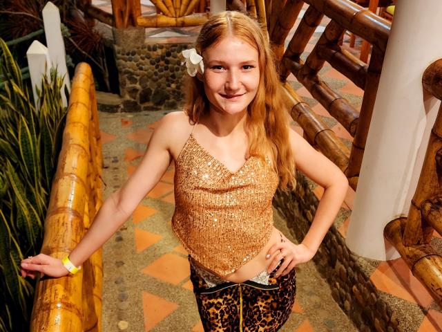 VanessaNorris, blonde, sparkly gold top, small tits, leopard pants, white flower in hair, smiling, hands on bamboo rail, yellow bracelet, standing on patterned tile.
