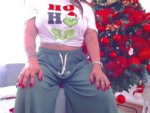 BelletinaXx with big tits, wearing a tied white shirt, green pants, red nails, sitting in front of a Christmas tree with red ornaments. Her hands are on her ass.