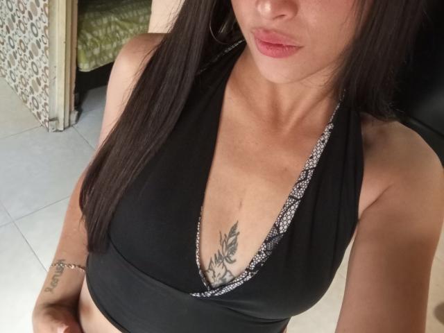 EmmyRouse with dark hair, wearing a black halter top, showing small tits and a tattoo of black leaves under her cleavage, with pink lips and a silver bracelet.