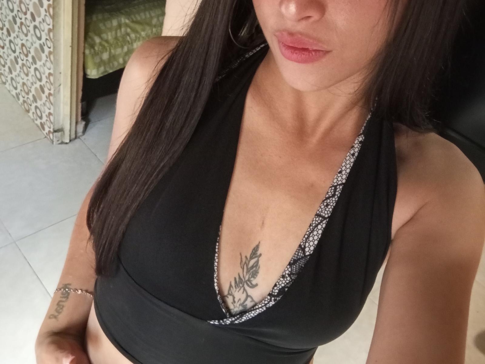 EmmyRouse with dark hair, wearing a black halter top, showing small tits and a tattoo of black leaves under her cleavage, with pink lips and a silver bracelet.