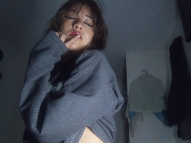 VanellopeLm with eyes closed, wearing a dark grey sweater, biting her finger, showing a small side of her bare ass, with messy brown hair and red lipstick.