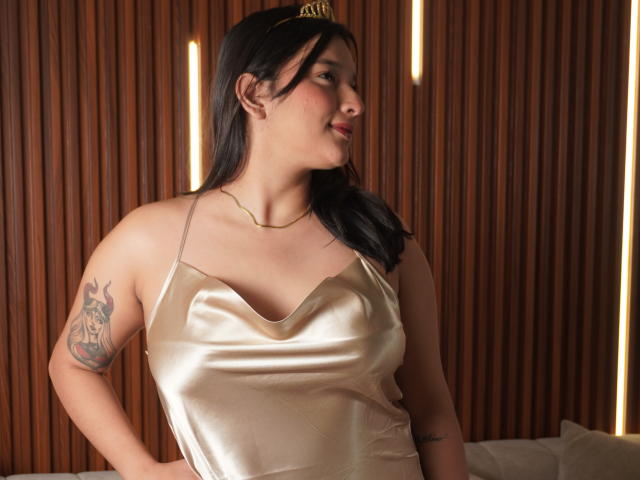 MiaEverly with tan satin dress, gold headband, black hair, small tits, tattoo on right arm, light skin, smiling, wooden wall background, standing, hands on hips, soft lighting.
