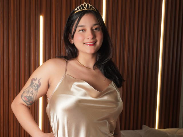 MiaEverly with dark hair, wearing a gold tiara and shiny satin top, smiling at the camera, showing a colorful tattoo on her left arm. Boobs visible.