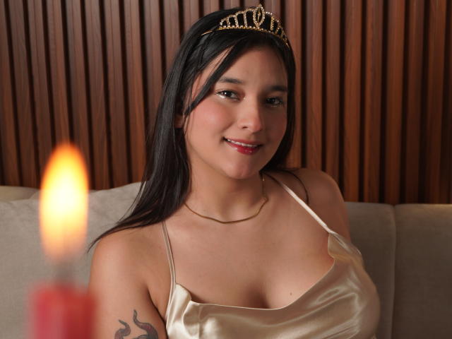 MiaEverly with black hair, wearing a gold tiara and shiny satin top, smiling, showing her tits, has a tattoo on her right arm, and a lit candle in front.