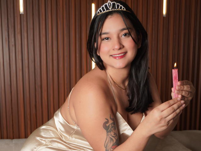 MiaEverly with black hair, wearing a silver tiara and shiny satin dress, holding a pink candle, smiling, tattoo on right arm, wood-paneled background, sitting on white surface.